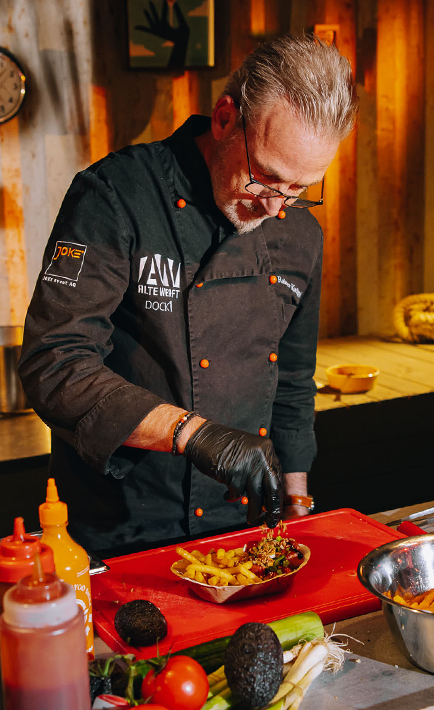 A man in a black chef's coat is preparing food in a kitchen. He is pouring something onto a bowl of food, which contains avocado.