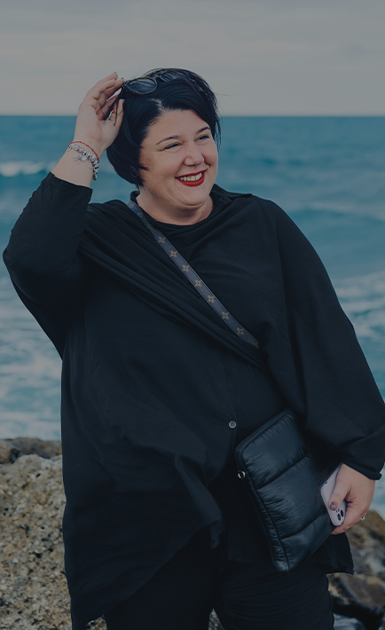 The image features a woman standing by the ocean, wearing a black outfit, and holding a purse. She is smiling and looking at the camera.