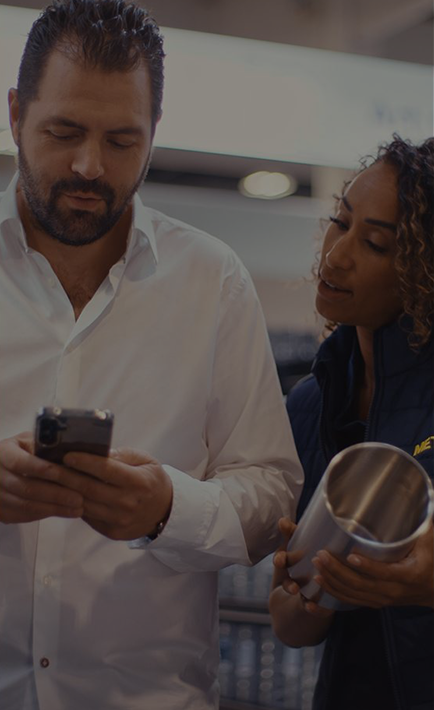 In the image, a man and a woman are standing next to each other. The woman is holding a cup, while the man is holding a cell phone. They seem to be engaged in a conversation or activity together.