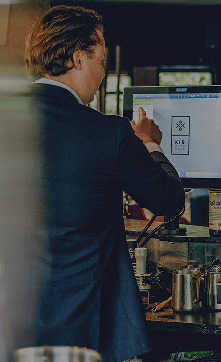 A man is standing at a counter with a screen, pointing at something on the screen.