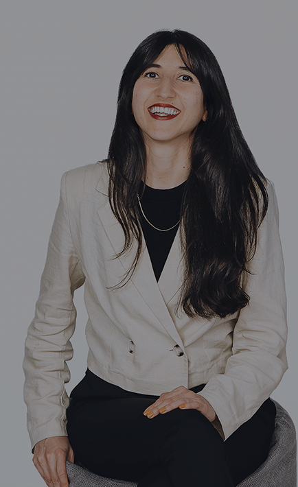 The image features a woman sitting on a chair, wearing a beige suit jacket and a black shirt. She has dark hair and is smiling, showing her teeth. The background is white, making her stand out in the picture.