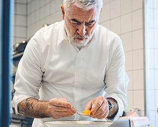 A cook ist standing in the kitchen preparing food with a spoon.